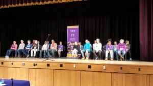 All 23 participants of the Spelling Bee sitting in chairs on the stage. 