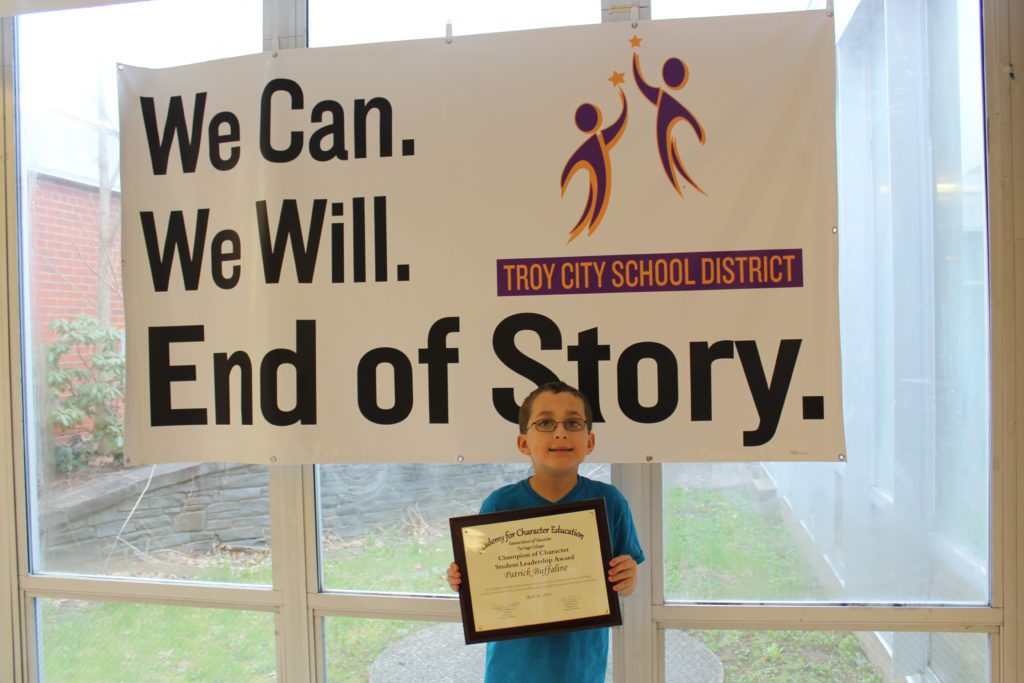 Patrick holding his plaque in front of the Troy CSD "We can, we will, end of story" banner.