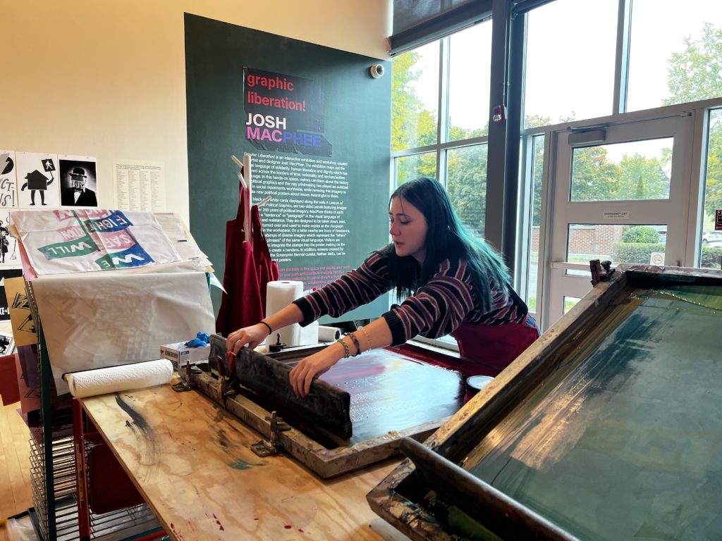 A student works at a screen printing machine in front of a wall of windows in an art gallery