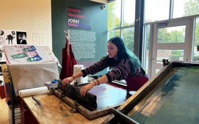 A student works at a screen printing machine in front of a wall of windows in an art gallery