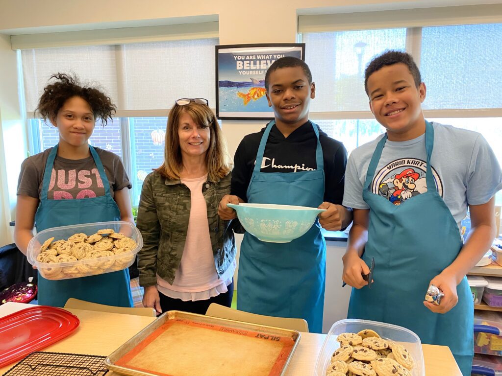 a teacherand three students hold bowls standing behind a counter in the kitchen.