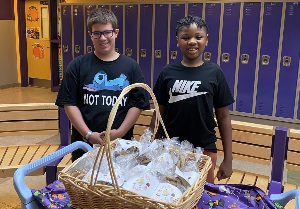 two students stand behind a rolling cart with a basket of cookies