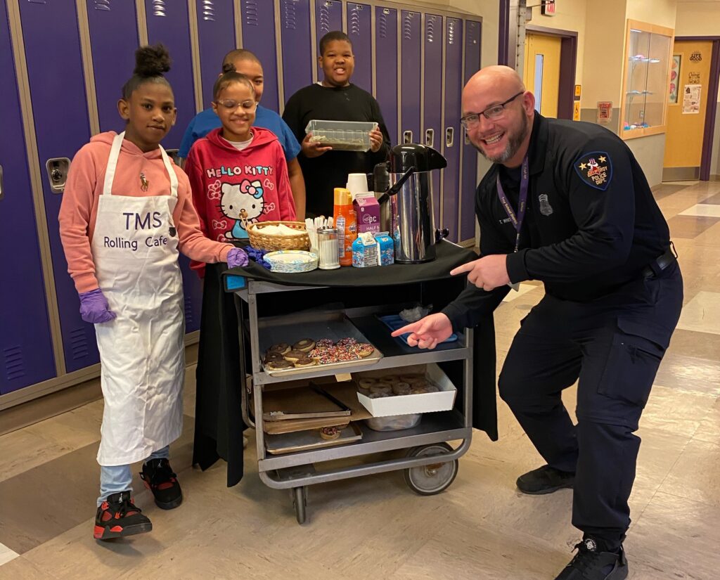 students pose with a school resource officer in front of the baking cart.