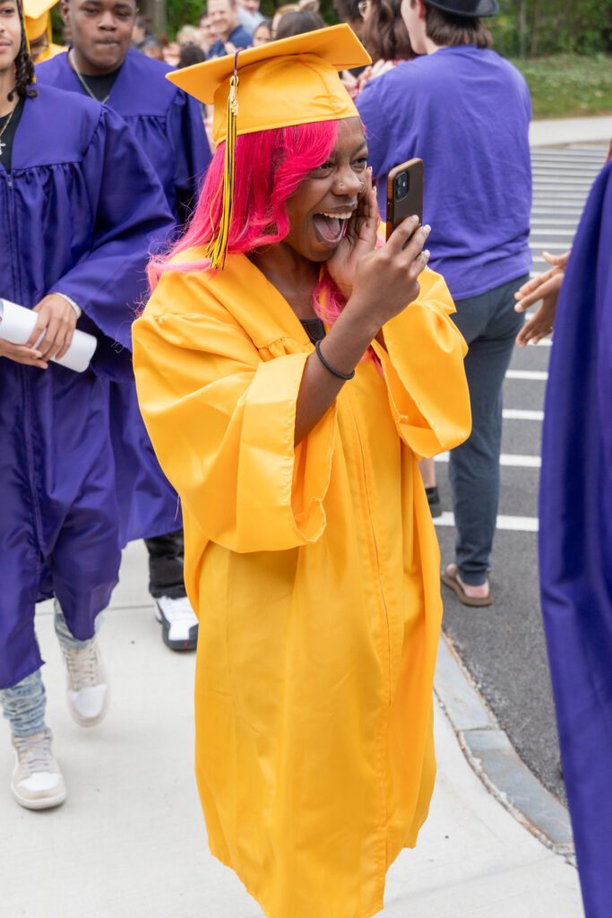 a female graduate in a yellow cap and gown records her entrance to graduation on her cell phone