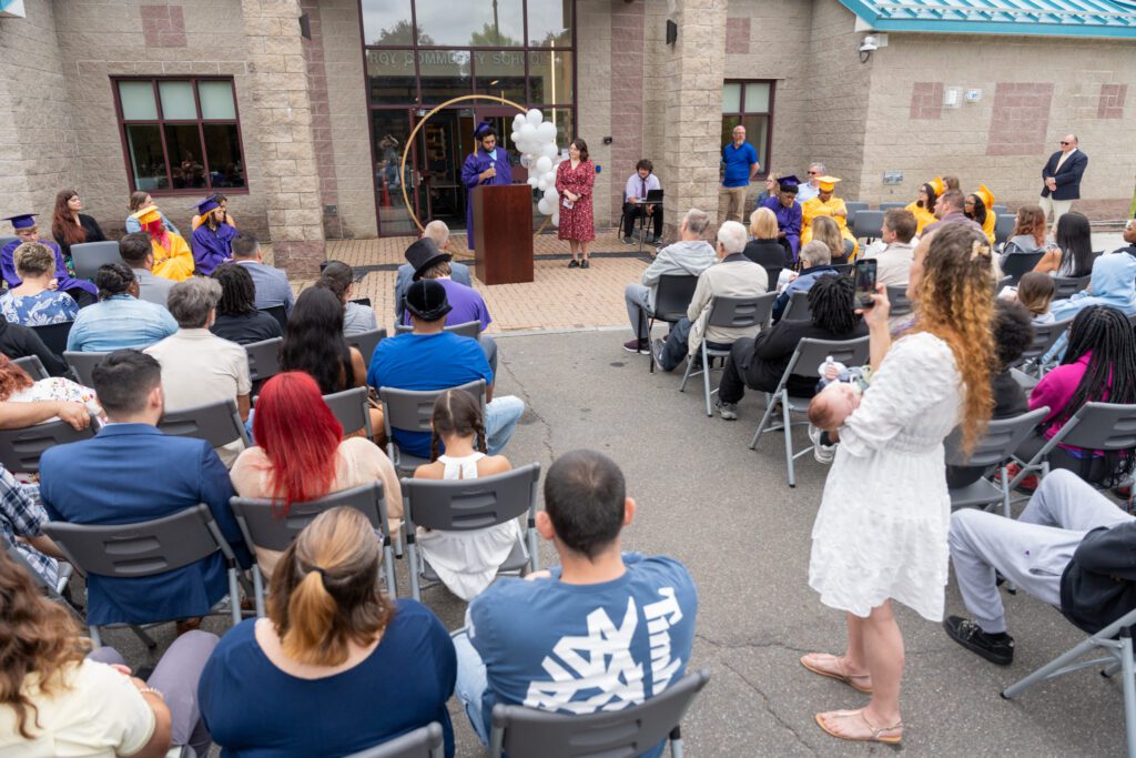 wide shot of graduates in front of a school with and audience seated in chairs facing them.