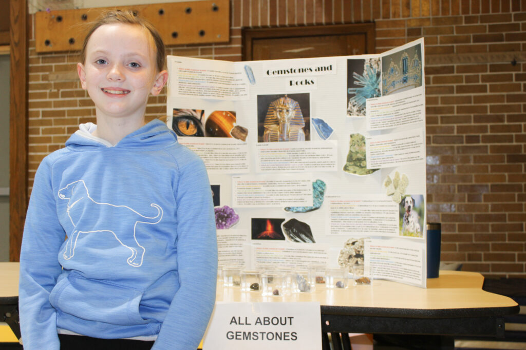 A young student smiles in front of her science fair project about gemstones and rocks, featuring a colorful display board and samples.