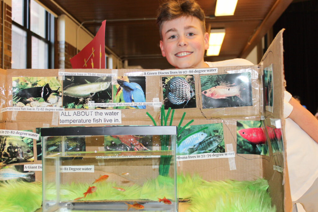 A student stands behind a science fair project about fish and water temperature, featuring a fish tank and display board with fish photos.