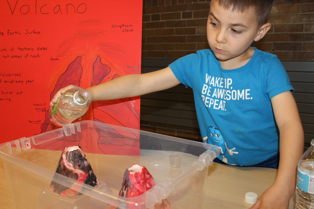 A young boy pours liquid into a model volcano during a school science fair. A red poster with a volcano diagram is displayed behind him.