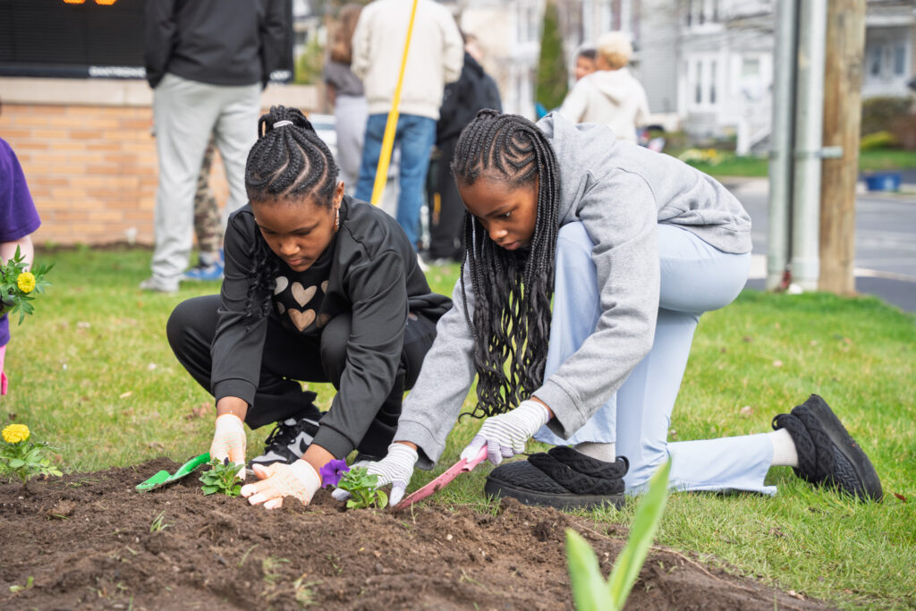 Two School 16 students carefully plant flowers in the front lawn during an Earth Day celebration, using gardening tools and gloves.
