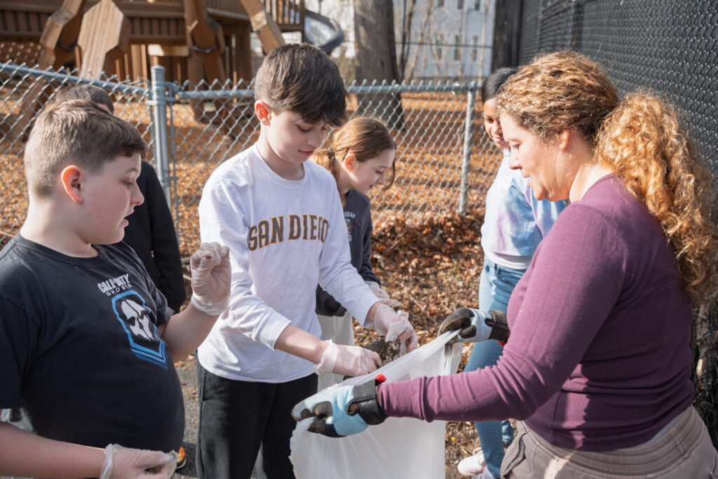 A group of School 16 students and an adult collect litter in a trash bag near the playground fence during the Earth Day clean-up.