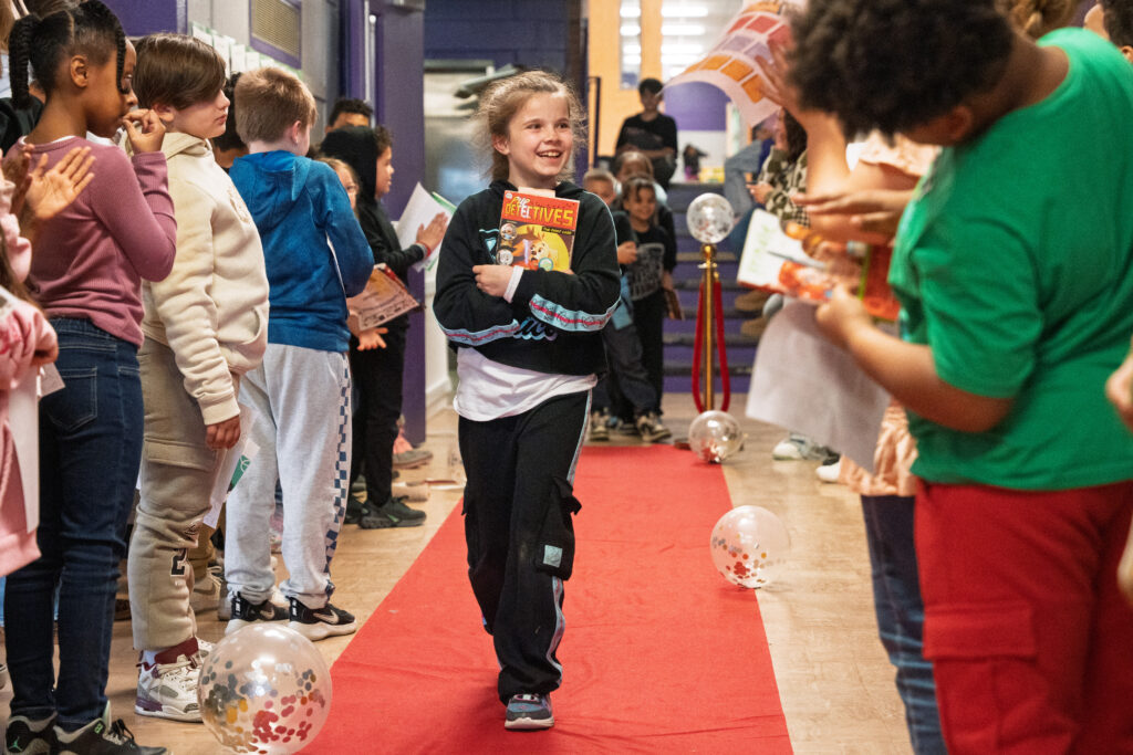 A smiling third-grade student walks down a red carpet holding a book, while classmates cheer along the hallway at Carroll Hill School.