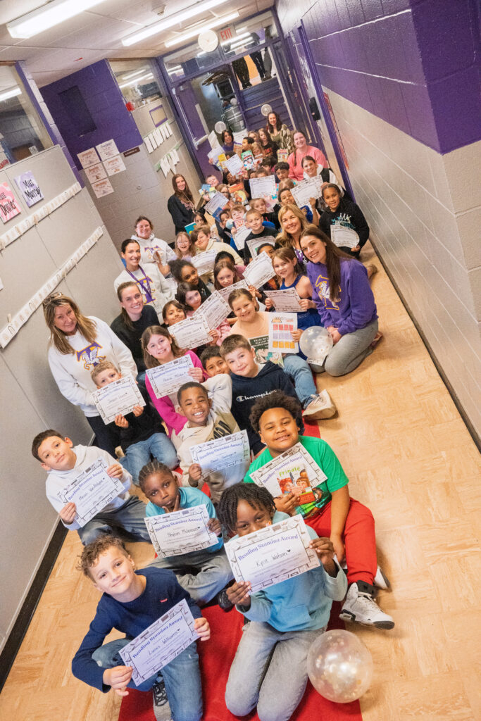A large group of Carroll Hill third-grade students and staff pose in a school hallway holding reading certificates and favorite books.