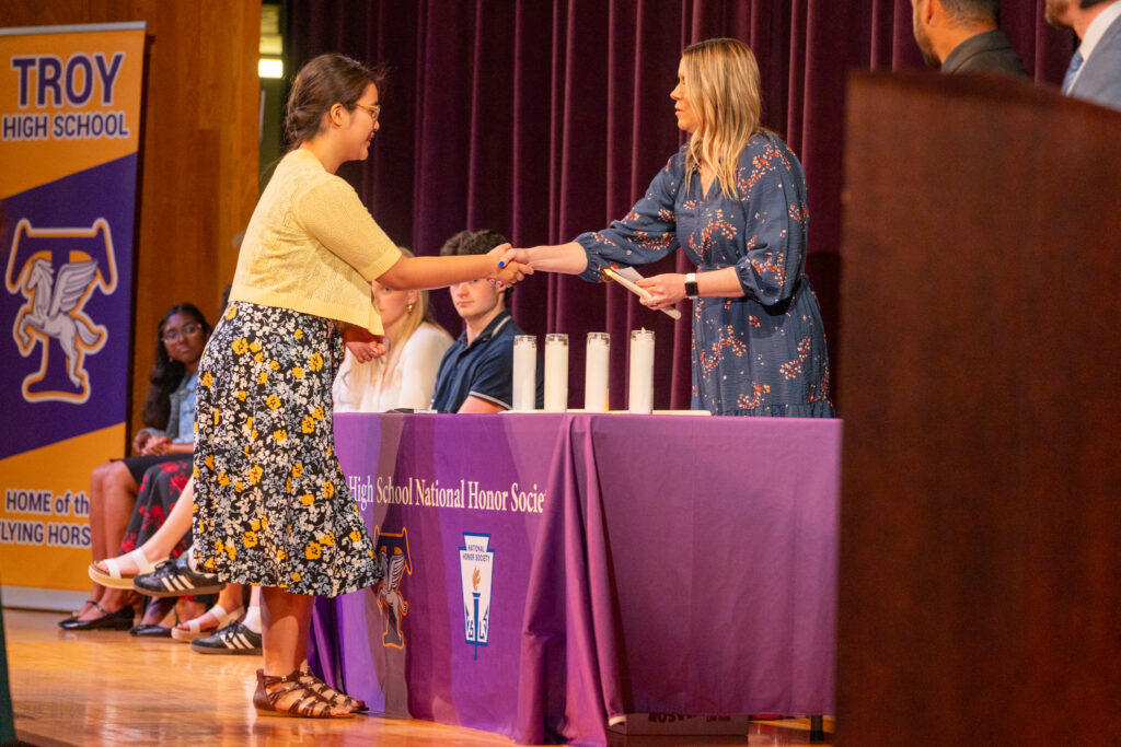 A student is inducted into the National Honor Society at Troy High School, shaking hands with a staff member while receiving a certificate on stage. Several students are seated behind a purple NHS table, with a Troy High School banner visible in the background.
