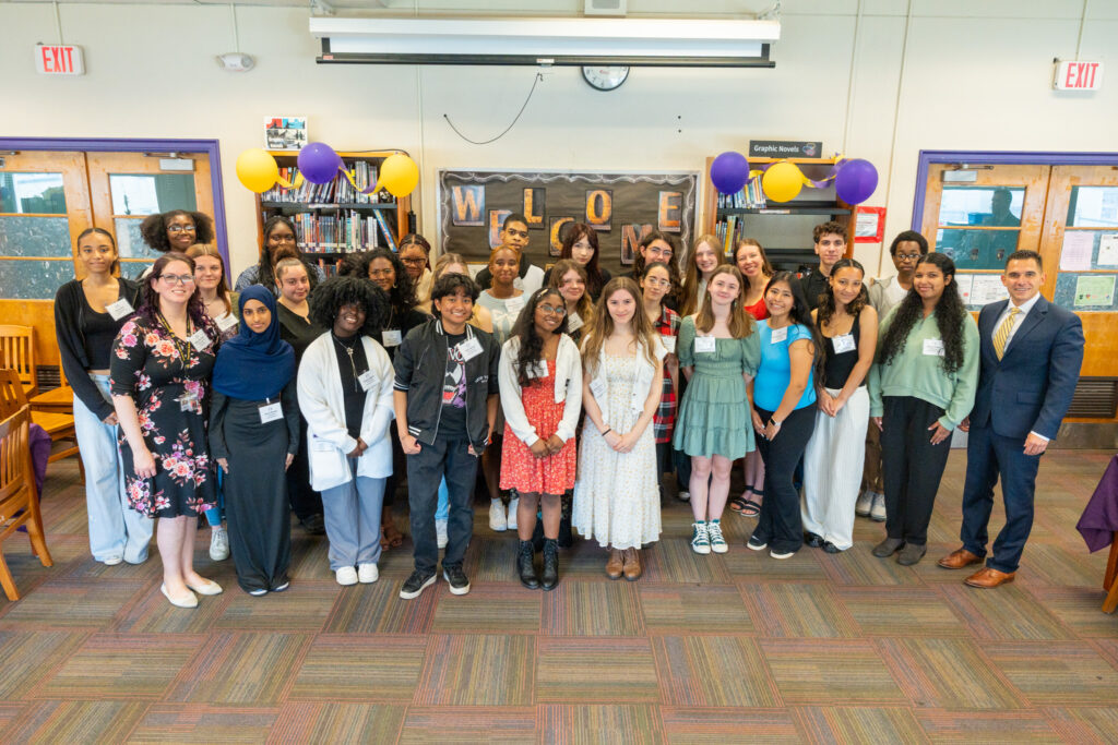 A large group of Troy High School students and staff pose together in the school library during a celebratory event, with a "Welcome" sign and purple and gold balloons in the background.