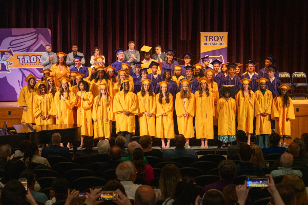A large group of high school graduates wearing yellow and purple caps and gowns stand on a stage in front of an audience during a graduation ceremony at Troy High School. A backdrop and banners display the school name and logo.