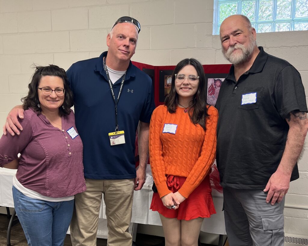 Four people stand together smiling for a photo in a community room. Behind them is a red tri-fold display board with photos and a white table. 