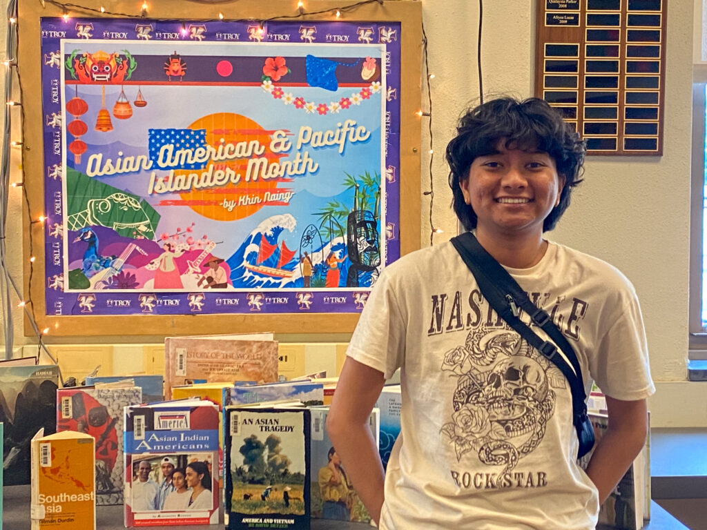 A student stands in front of a decorated library display celebrating Asian American & Pacific Islander Month, featuring colorful cultural artwork and a table of related books.