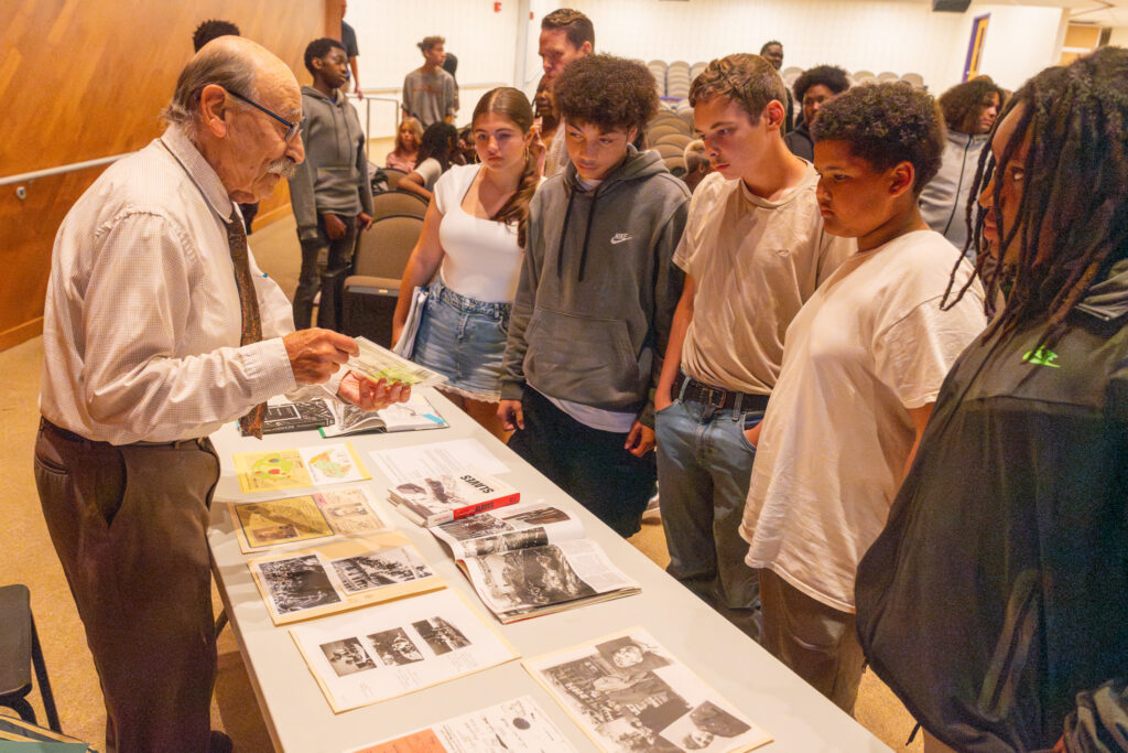 Holocaust survivor Ivan Vamos speaks to a group of Troy Middle School eighth grade students, showing historical documents spread across a table.