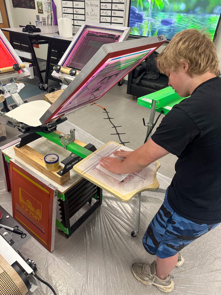 Student pressing a design onto paper at a screen printing station in an art classroom, surrounded by colorful screens and equipment.