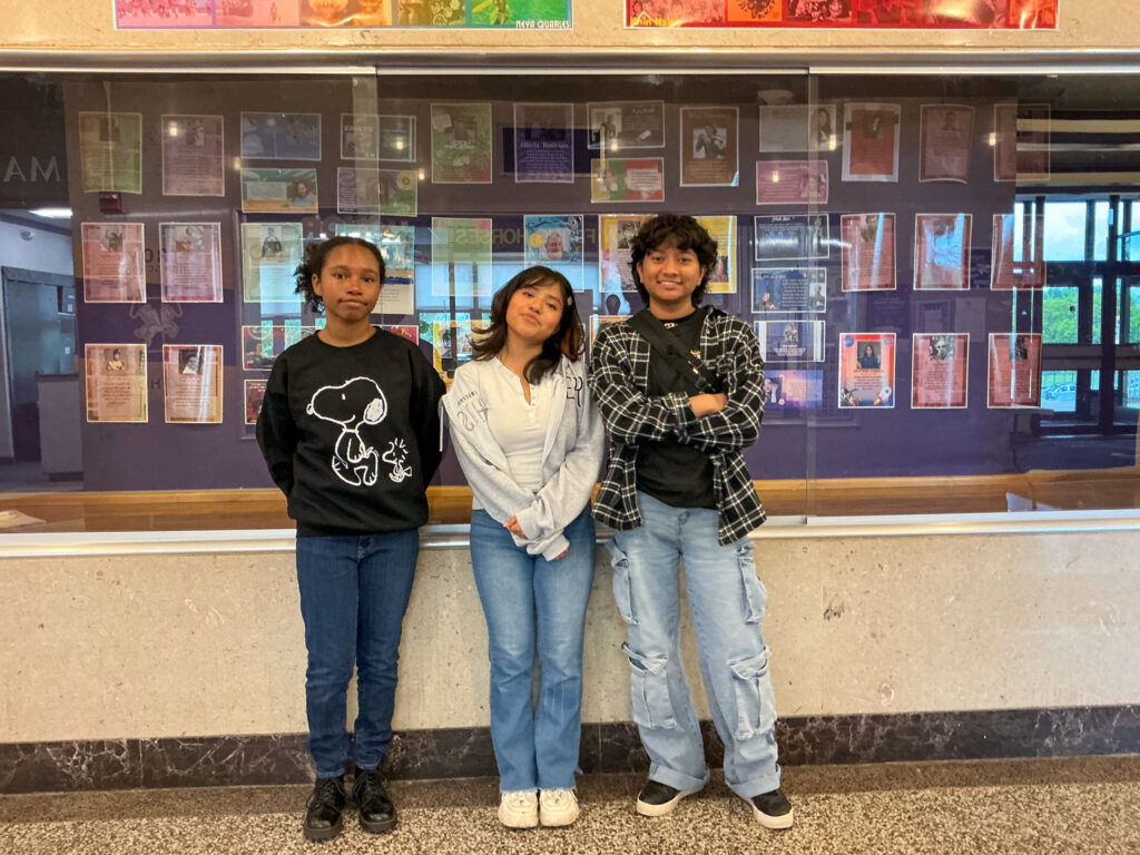 Three students stand together in front of a large hallway display case featuring educational posters and tributes for Asian American & Pacific Islander Month.
