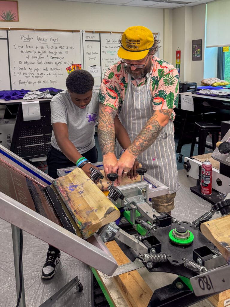 Ralph Stollenwerk and student collaborating on a screen printing project in a classroom, pressing ink onto a shirt using a screen printing press.
