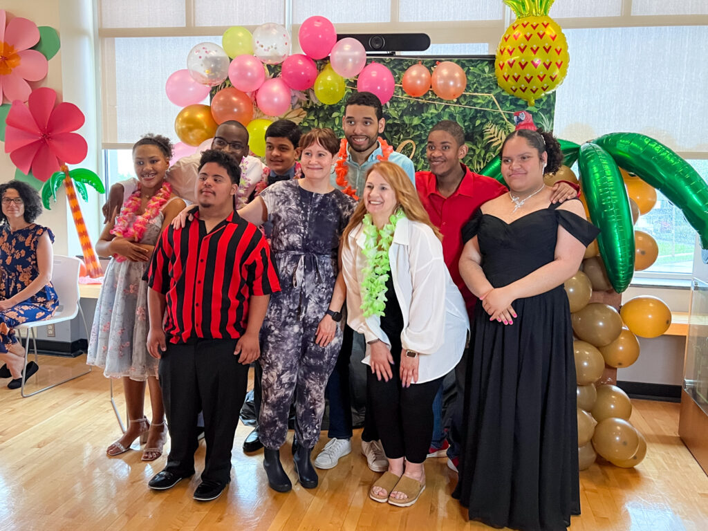 Group of Life Skills students and staff smiling together in front of a colorful balloon display with tropical decorations, including a pineapple and palm tree.