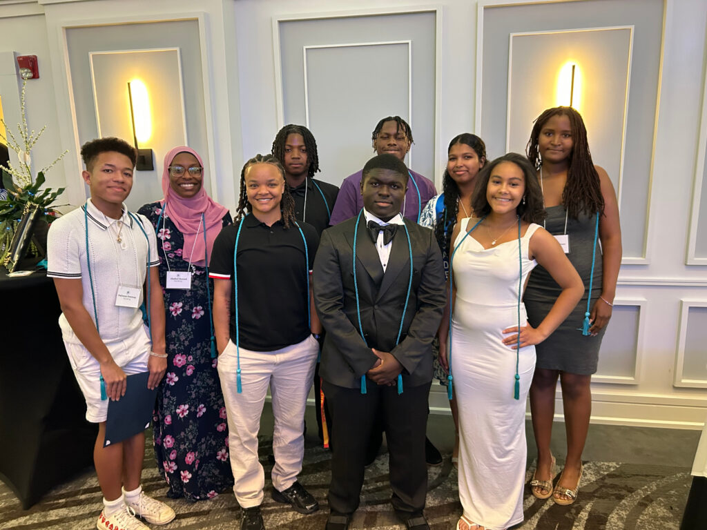 Nine Troy High School seniors smiling and standing in two rows at the 518 ElevatED Senior Scholar Celebration. They are wearing honor cords and name tags in a well-lit event space with white paneled walls.