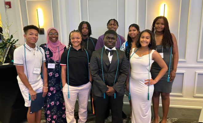 Nine Troy High School seniors smiling and standing in two rows at the 518 ElevatED Senior Scholar Celebration. They are wearing honor cords and name tags in a well-lit event space with white paneled walls.