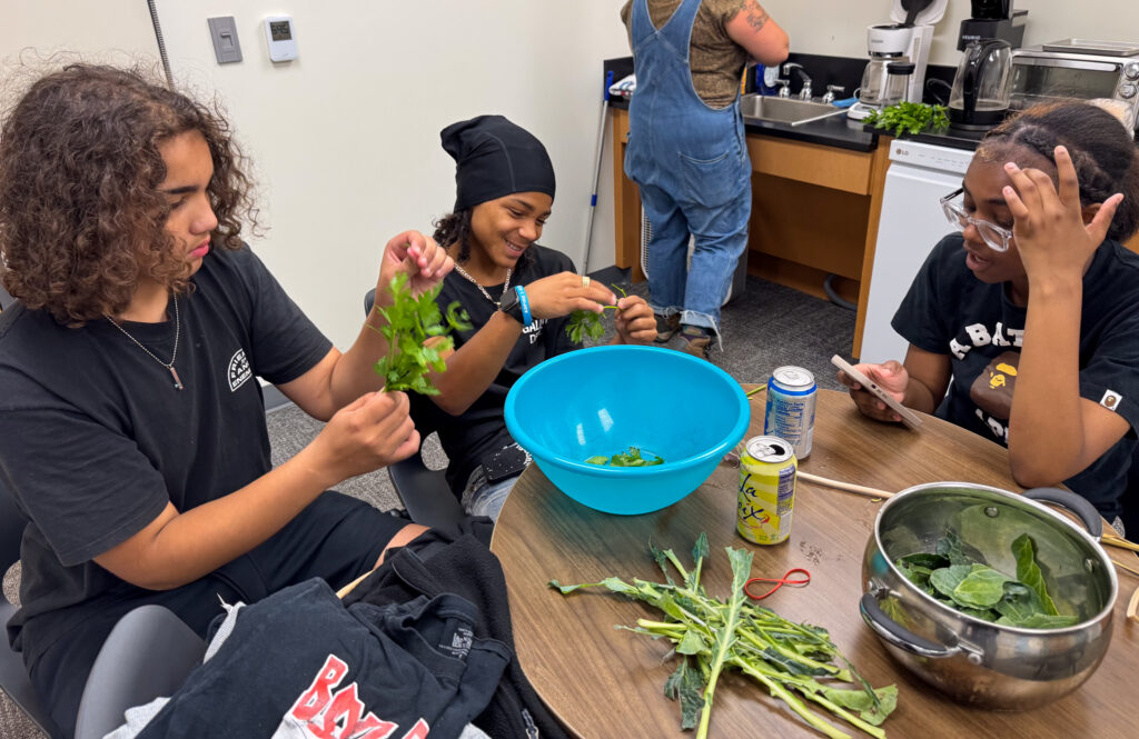 Three Troy Community School students sit around a table preparing leafy greens during the RISE Summer Youth Employment program. They are smiling and working together, with a large blue mixing bowl and a pot of greens on the table.