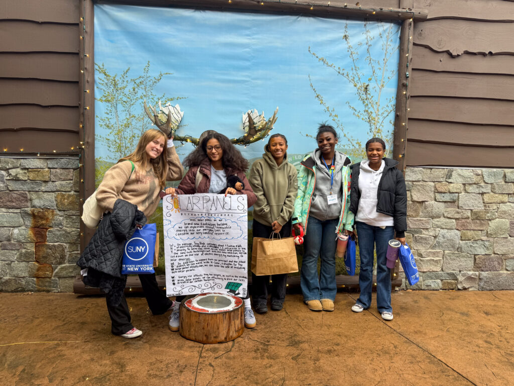 Five Troy High School students stand together outdoors in front of a backdrop featuring trees and a blue sky. They are smiling and holding bags from the event. Two students in the center hold a large poster titled “Solar Panels” that outlines their climate action project.