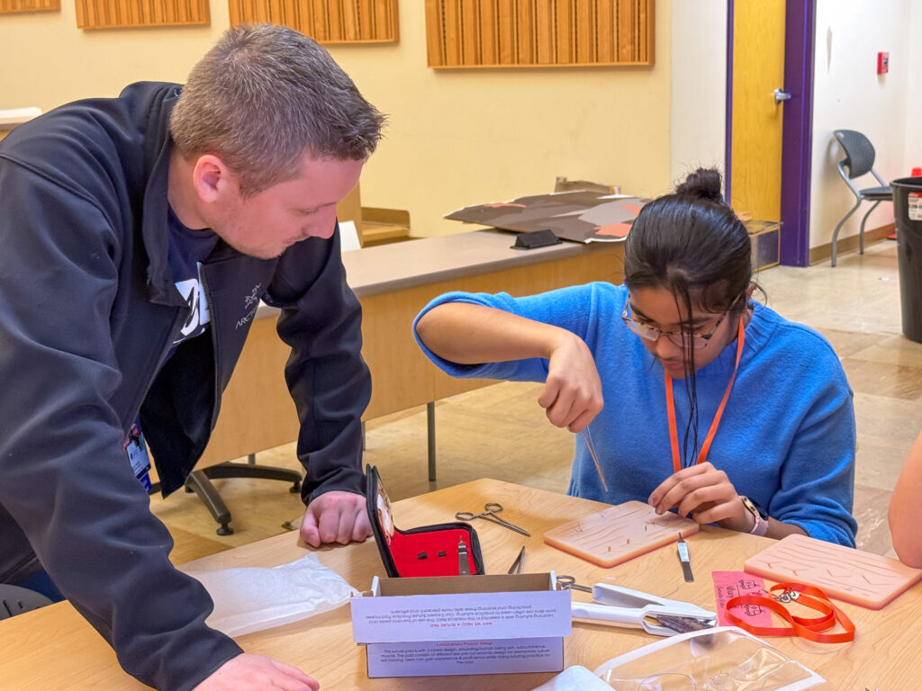 A female student sitting at a table practices suturing on a synthetic suture pad while an adult looks on.