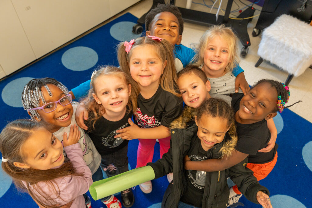 group of kindergarten students standing together on a rug, looking up at the camera and smiling