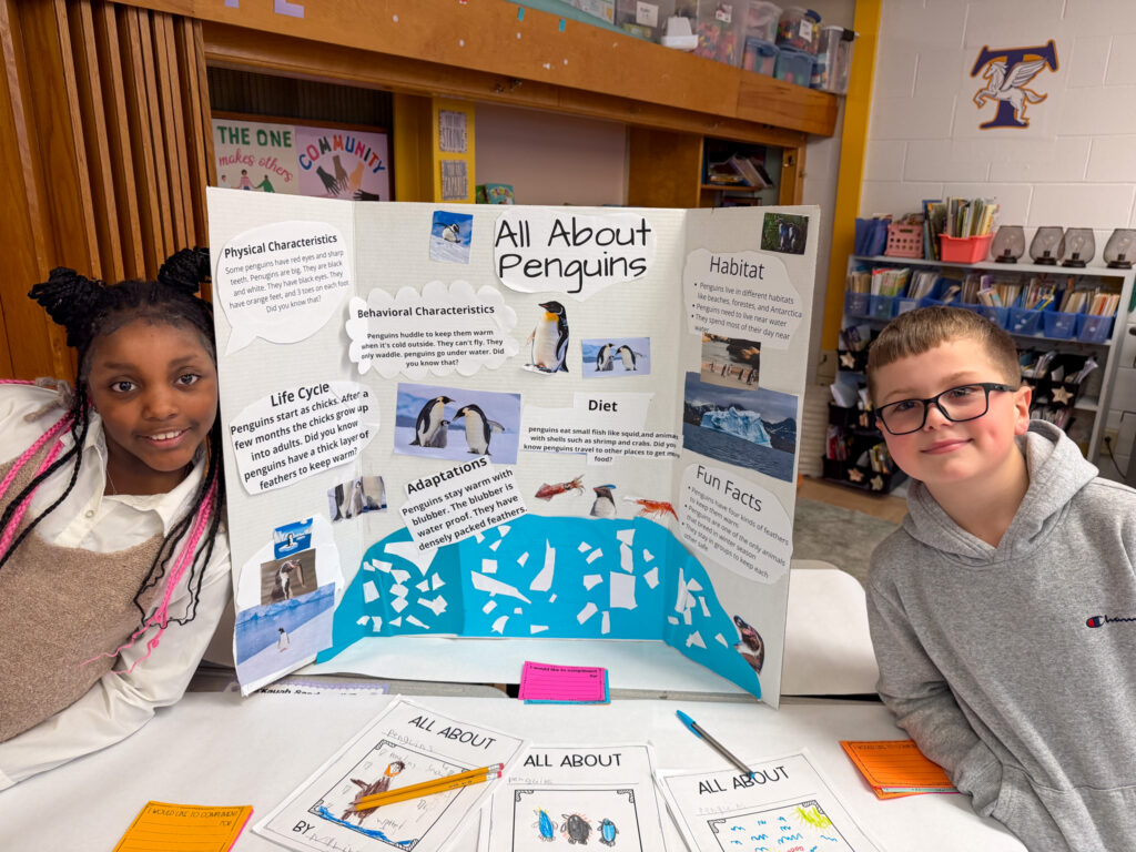two students smile next to their "all about penguins" poster in a classroom at Carroll Hill School