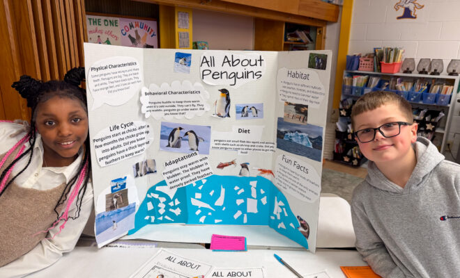 two students smile next to their "all about penguins" poster in a classroom at Carroll Hill School